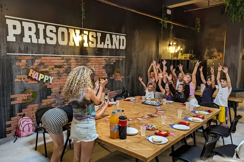 Des enfants attablés lèvent les bras devant un gâteau, sous l'enseigne Prison Island